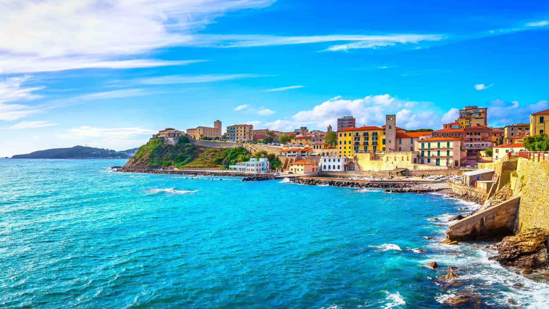 A panoramic view of the colorful, historic town of Piombino in Tuscany, Italy, with buildings perched on a cliffside overlooking the stunning turquoise waters of the Ligurian Sea.