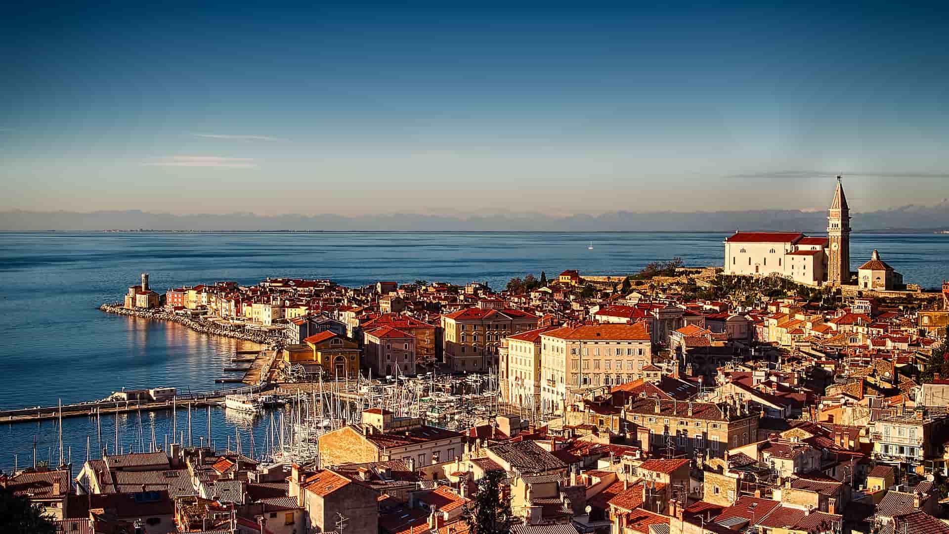 A scenic, high-angle view of the historic coastal town of Piran, Slovenia, with its densely packed terracotta roofs, a marina filled with boats, and a church with a bell tower overlooking the Adriatic Sea at sunset.