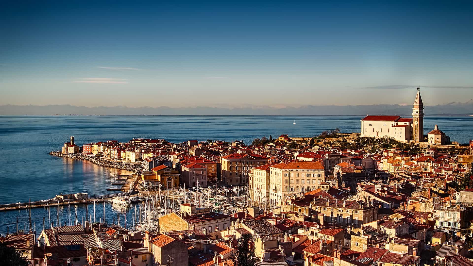 A scenic, high-angle view of the historic coastal town of Piran, Slovenia, with its densely packed terracotta roofs, a marina filled with boats, and a church with a bell tower overlooking the Adriatic Sea at sunset.