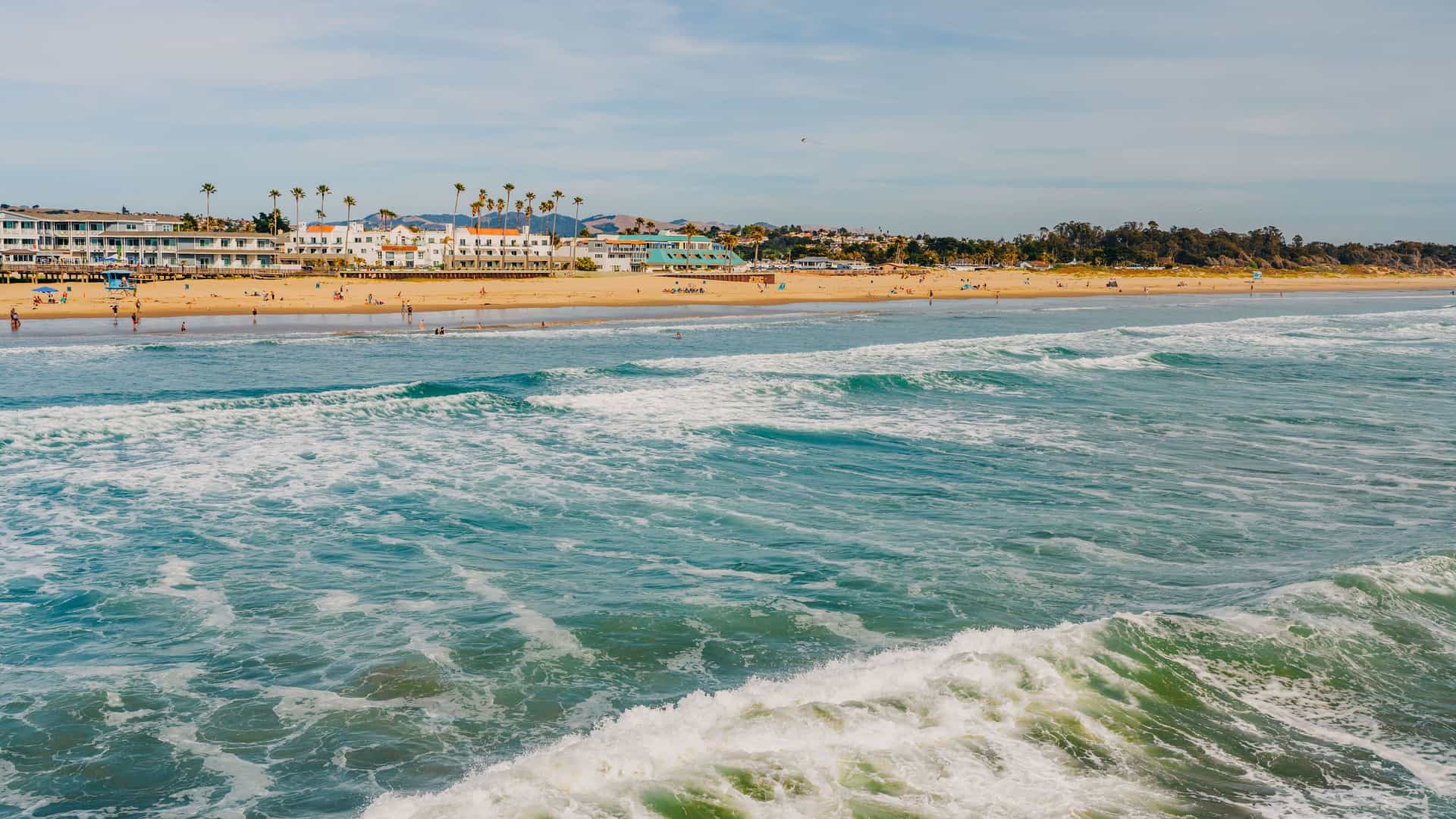 A horizontal view of Pismo Beach, California, with large blue-green waves rolling towards the shore. The sandy beach is populated with people, and a line of white buildings with red-tiled roofs and palm trees sits just beyond the sand. The sky is partly cloudy.