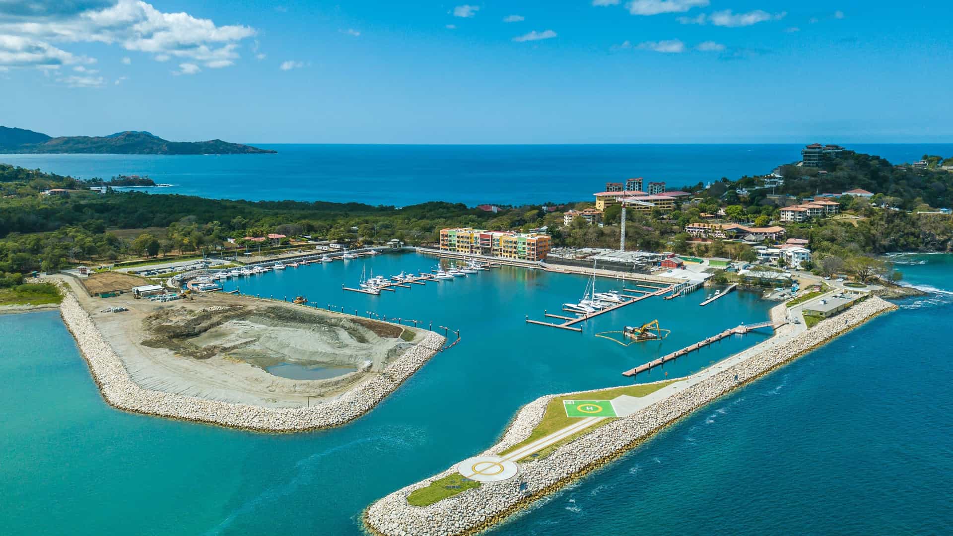 An aerial view of the marina at Playa Flamingo in Costa Rica. The turquoise harbor is filled with docked boats and is protected by two stone breakwaters, one of which has a helipad. The surrounding green hills are dotted with colorful buildings and houses, while the open ocean and a distant shoreline are visible in the background.