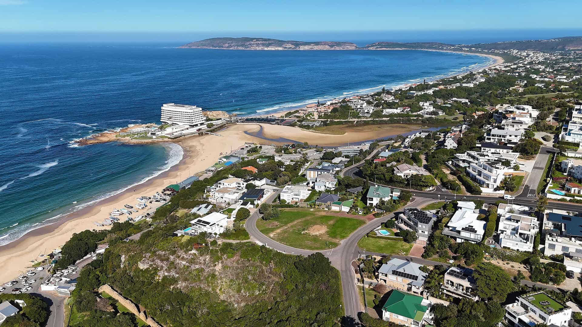 An aerial view of Plettenberg Bay, South Africa, showing a sweeping coastline with a wide sandy beach. The town's white houses and buildings are nestled on the green hills that slope down to the bay, where a river empties into the ocean. A prominent headland is visible in the distance under a clear blue sky.