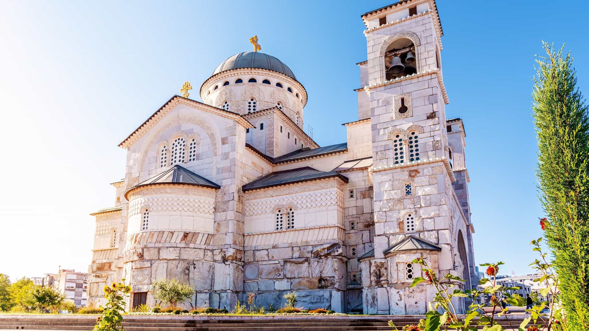 A close-up view of the Resurrection of Christ Orthodox Cathedral in Podgorica, Montenegro, highlighting its intricate white stone facade, domed roof, and a separate bell tower under a clear blue sky.