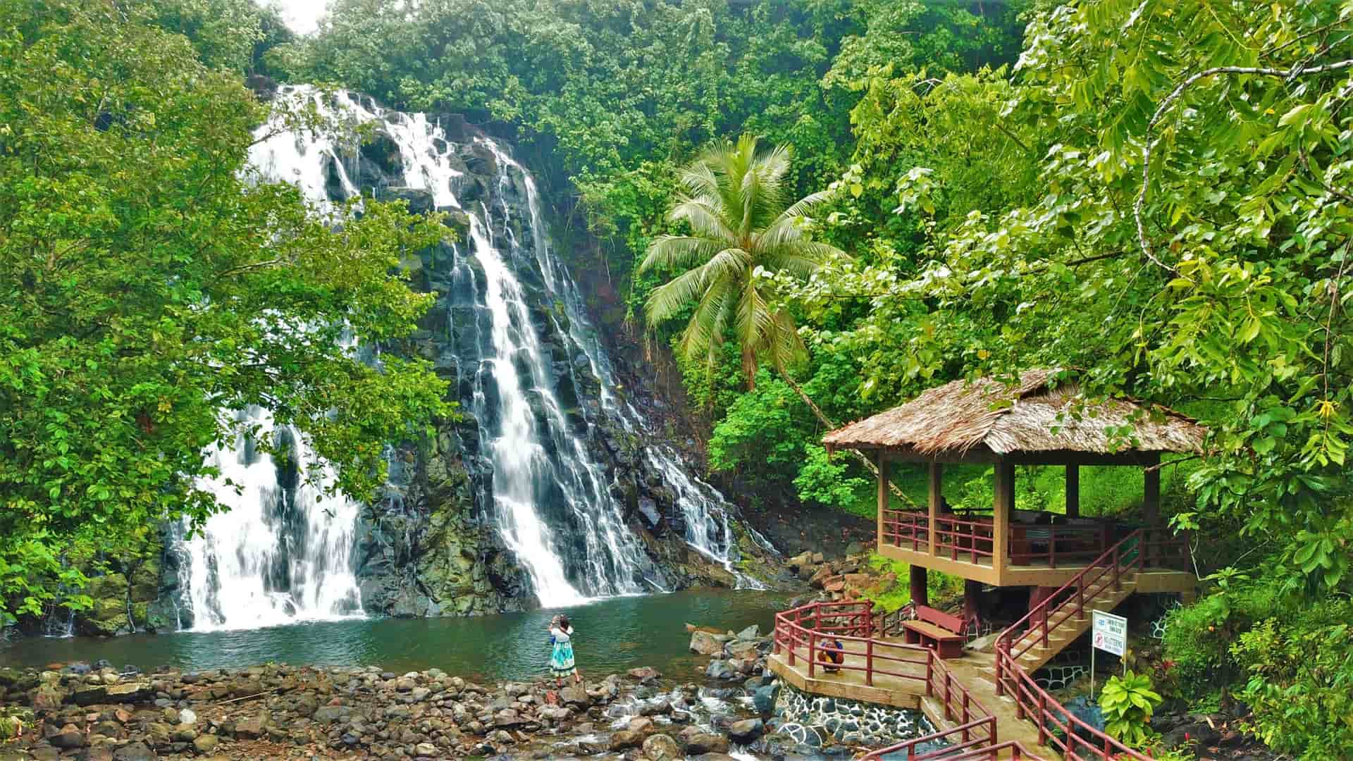 A stunning jungle waterfall cascades over black rocks, with a thatched-roof pavilion and palm trees on Pohnpei, Micronesia.