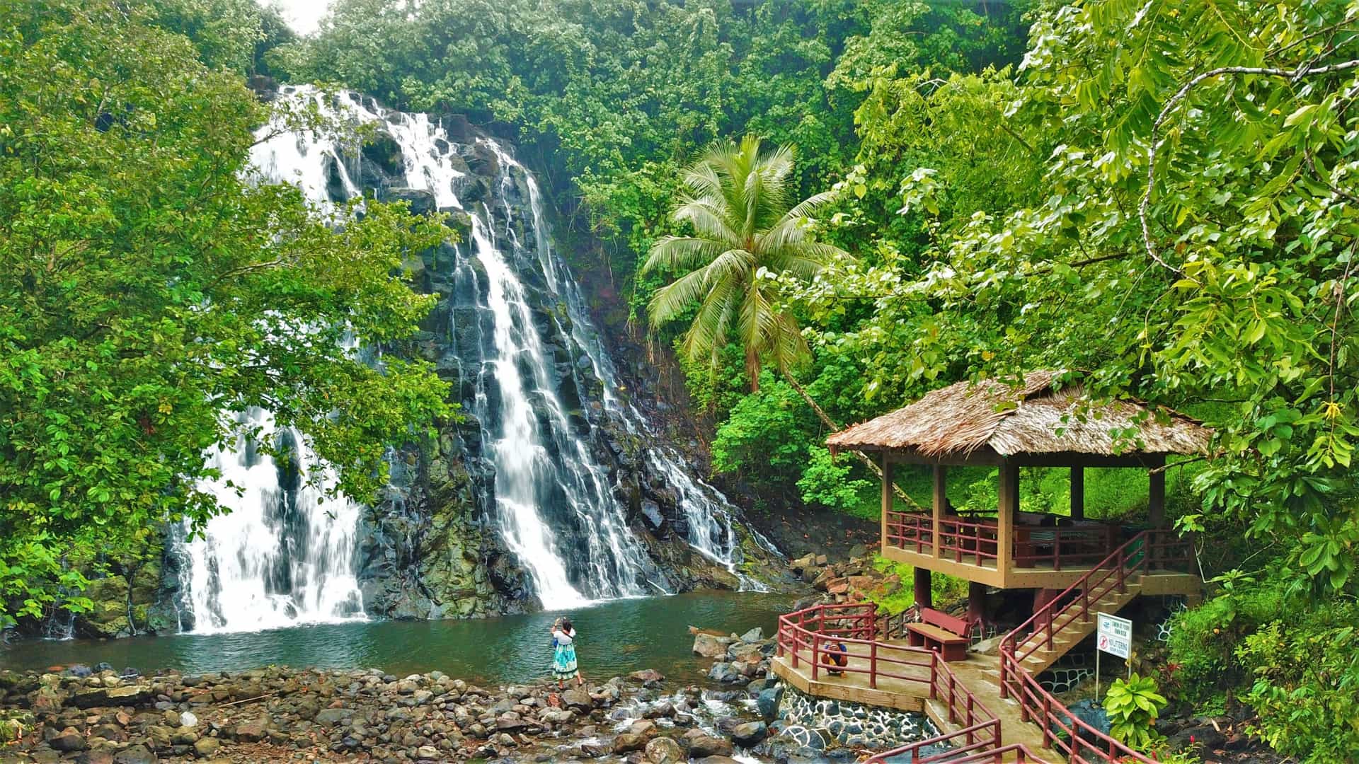 A stunning jungle waterfall cascades over black rocks, with a thatched-roof pavilion and palm trees on Pohnpei, Micronesia.