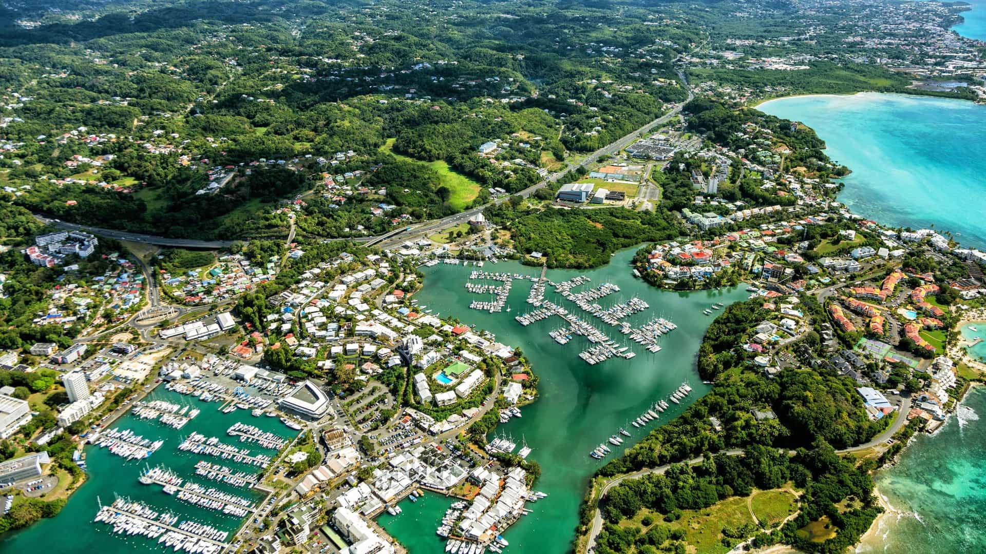 An aerial view of the vibrant marina and coastline of Pointe-à-Pitre, Guadeloupe, showing numerous boats docked in the harbor, surrounded by lush green hills and the turquoise Caribbean Sea.