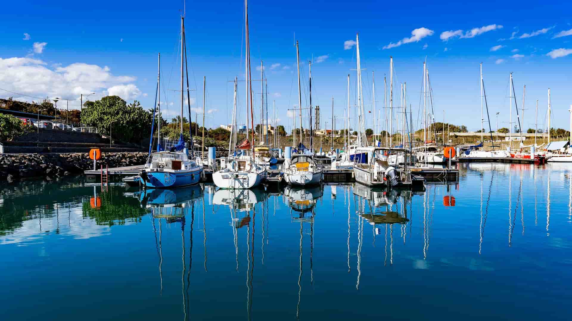 A scenic view of the marina at Pointe des Galets on Réunion Island, showing sailboats and yachts docked in the calm blue water with their reflections visible, under a clear sky.