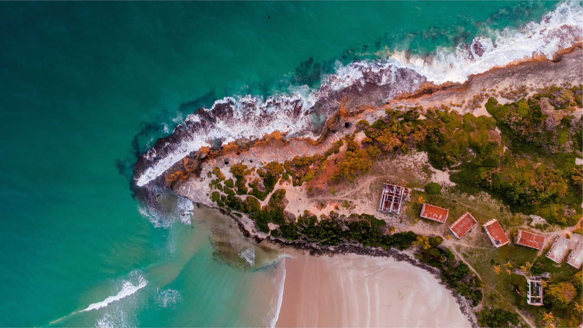 An aerial drone shot of the pristine coastline of Pomene National Reserve in Mozambique, showing a narrow, sandy peninsula dividing the bright turquoise waters of the Indian Ocean, with a stretch of green vegetation along the beach.