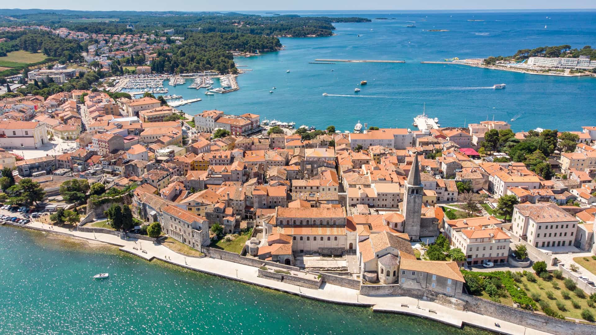 An aerial drone photograph of the historic old town of Poreč, Croatia, situated on a peninsula, showcasing its dense cluster of buildings with red-tiled roofs extending into the brilliant turquoise and blue water of the Adriatic Sea.