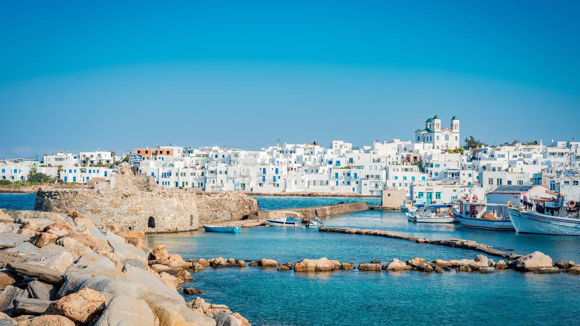 A bright, sunlit view of the coastal town of Poros, Greece, with its traditional white buildings and a prominent domed church on a hill. In the foreground, the remains of an old stone fortress sit in the turquoise water of a small harbor, where several boats are docked.