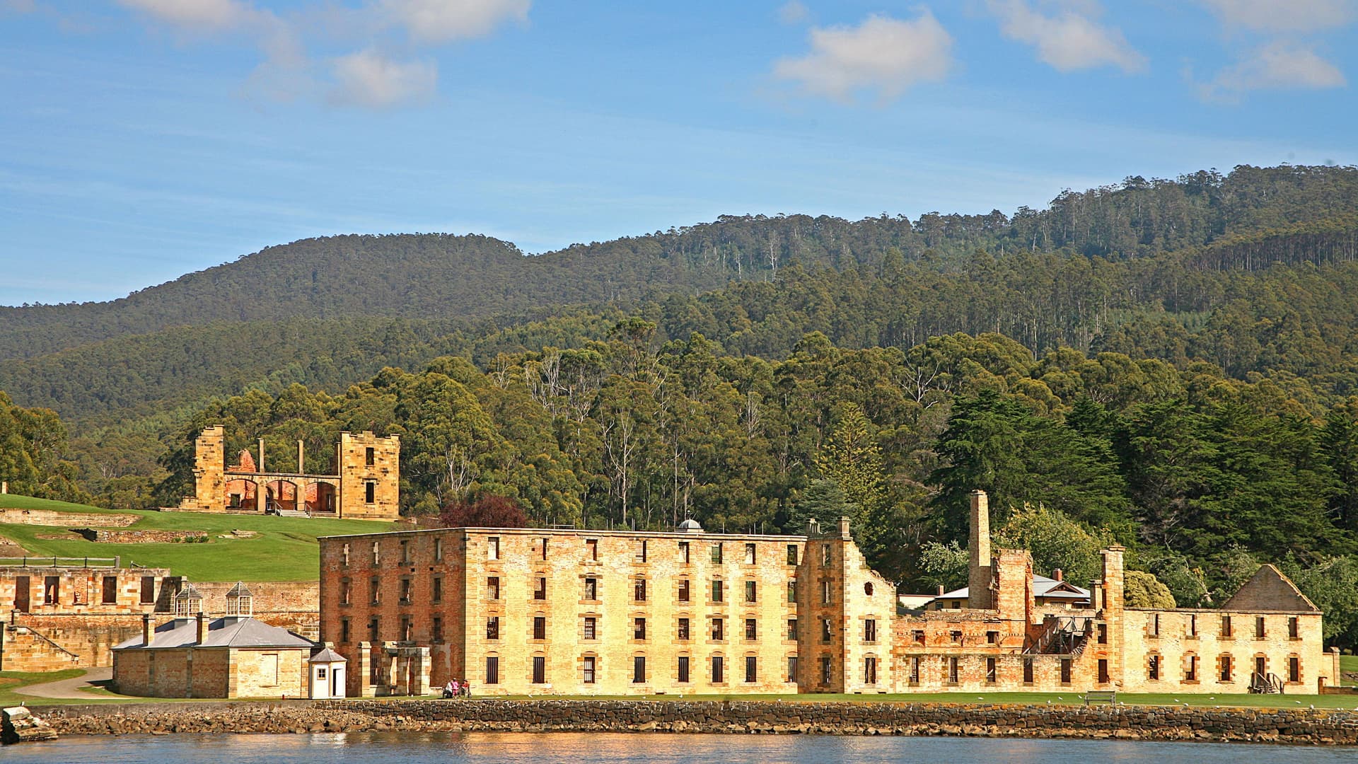 A panoramic view of the historic Port Arthur penal colony ruins in Tasmania, Australia, with multiple stone buildings and structures facing the water and a lush, tree-covered hillside in the background.