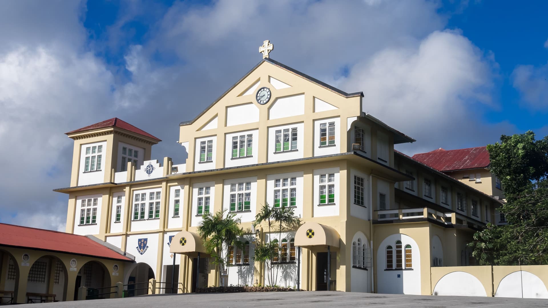 A classic, two-story colonial building, the Abbey of Our Lady of Exile, with a red roof, a clock tower, and arches in Port of Spain, Trinidad and Tobago, under a cloudy blue sky.