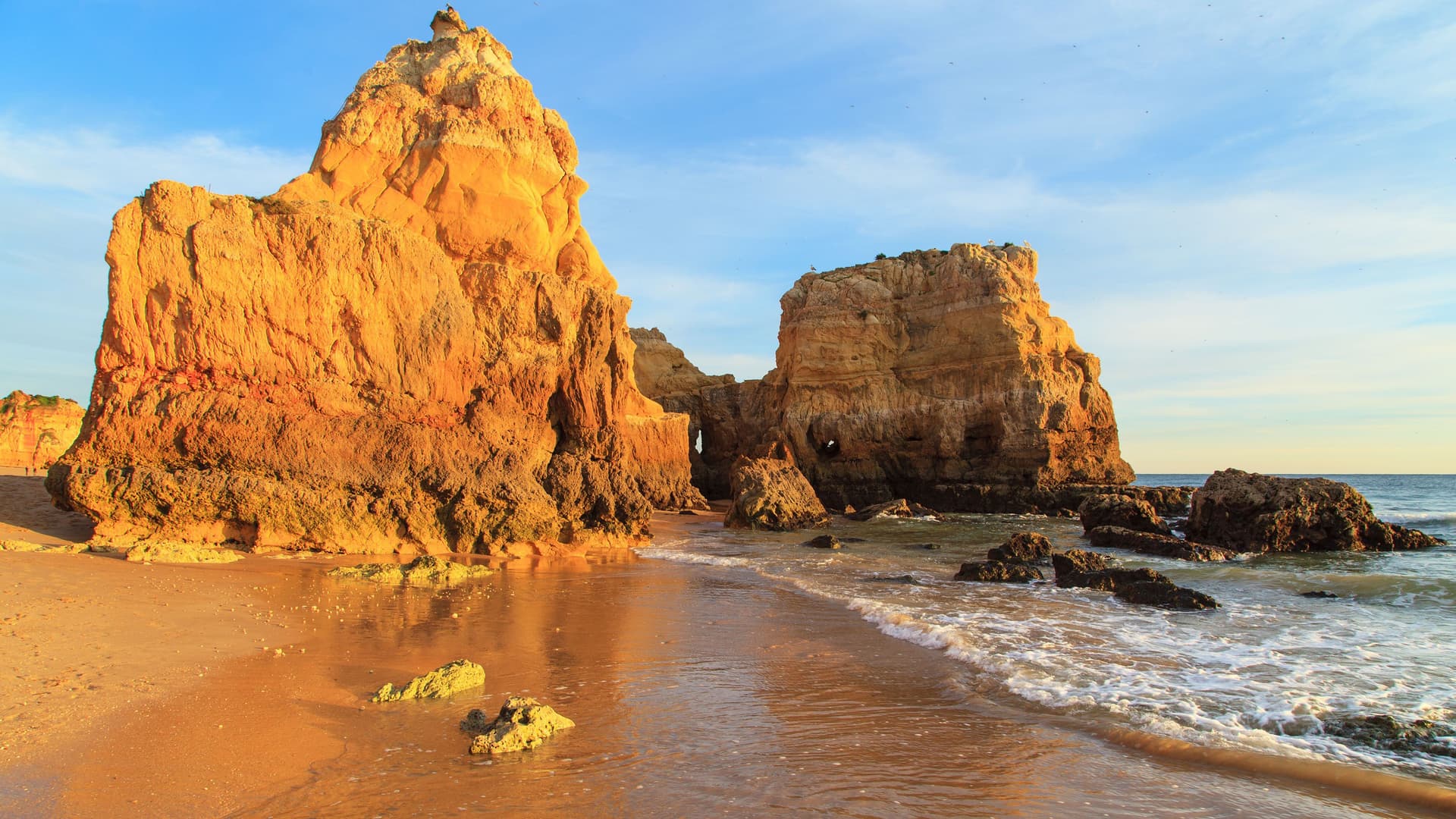 A golden-hour shot of a sandy beach in Portimão, Portugal, with the clear water of the Atlantic Ocean gently washing over the sand and the large, jagged limestone rock formations of the Algarve coast lit by the sun.