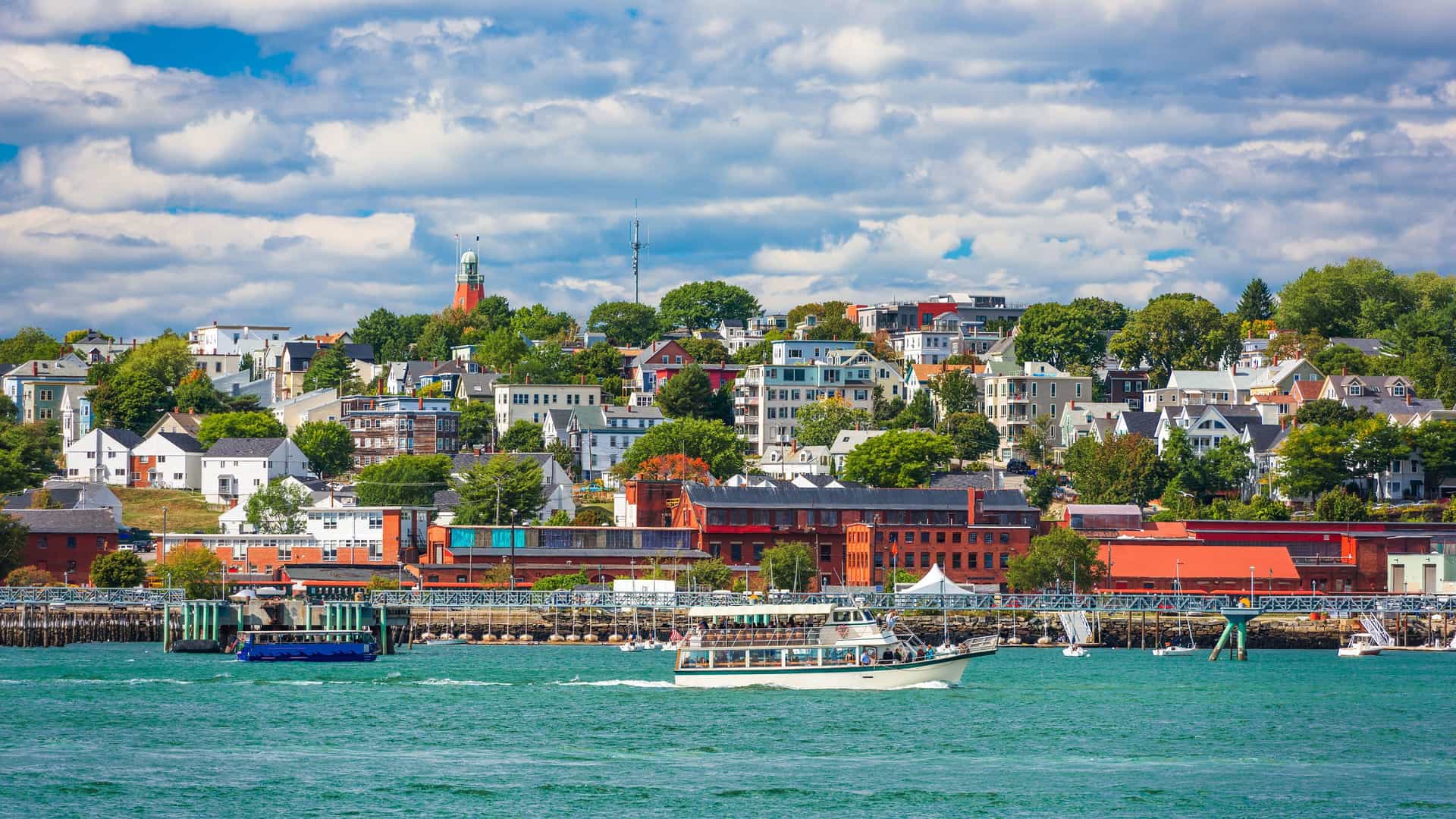 A panoramic view of the city of Portland, Maine, with a colorful waterfront and a variety of boats sailing on the water in the foreground and the city buildings and a lighthouse on the hill in the background.