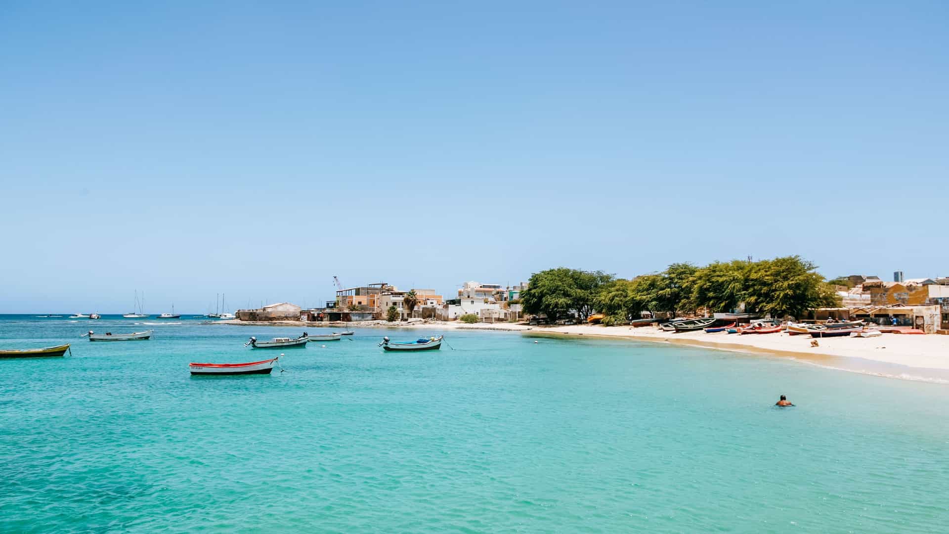A serene panoramic view of the Porto Antigo beach with its turquoise water, small fishing boats, and a few people swimming near the white sandy shore.
