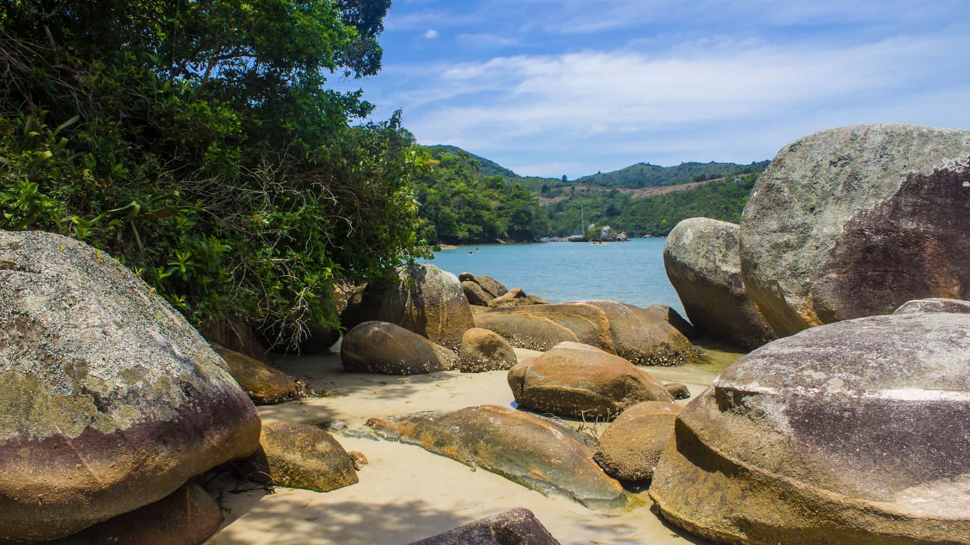 A tranquil beach at Porto Belo, Brazil, featuring a beautiful bay with calm blue water, lush green vegetation, and large, smooth boulders on the shoreline.