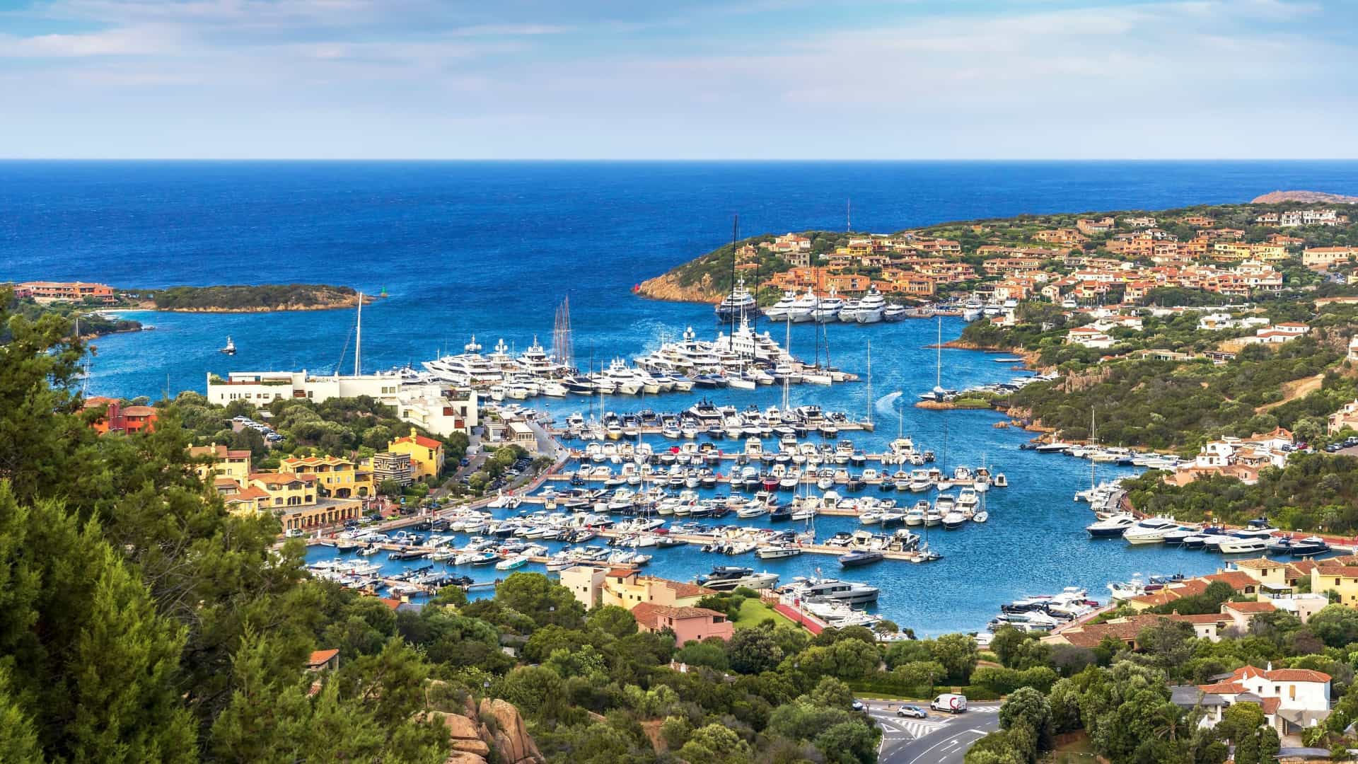 A high-angle view of the bustling Porto Cervo marina on Sardinia's Costa Smeralda, filled with luxury yachts and surrounded by coastal homes and lush Mediterranean vegetation.