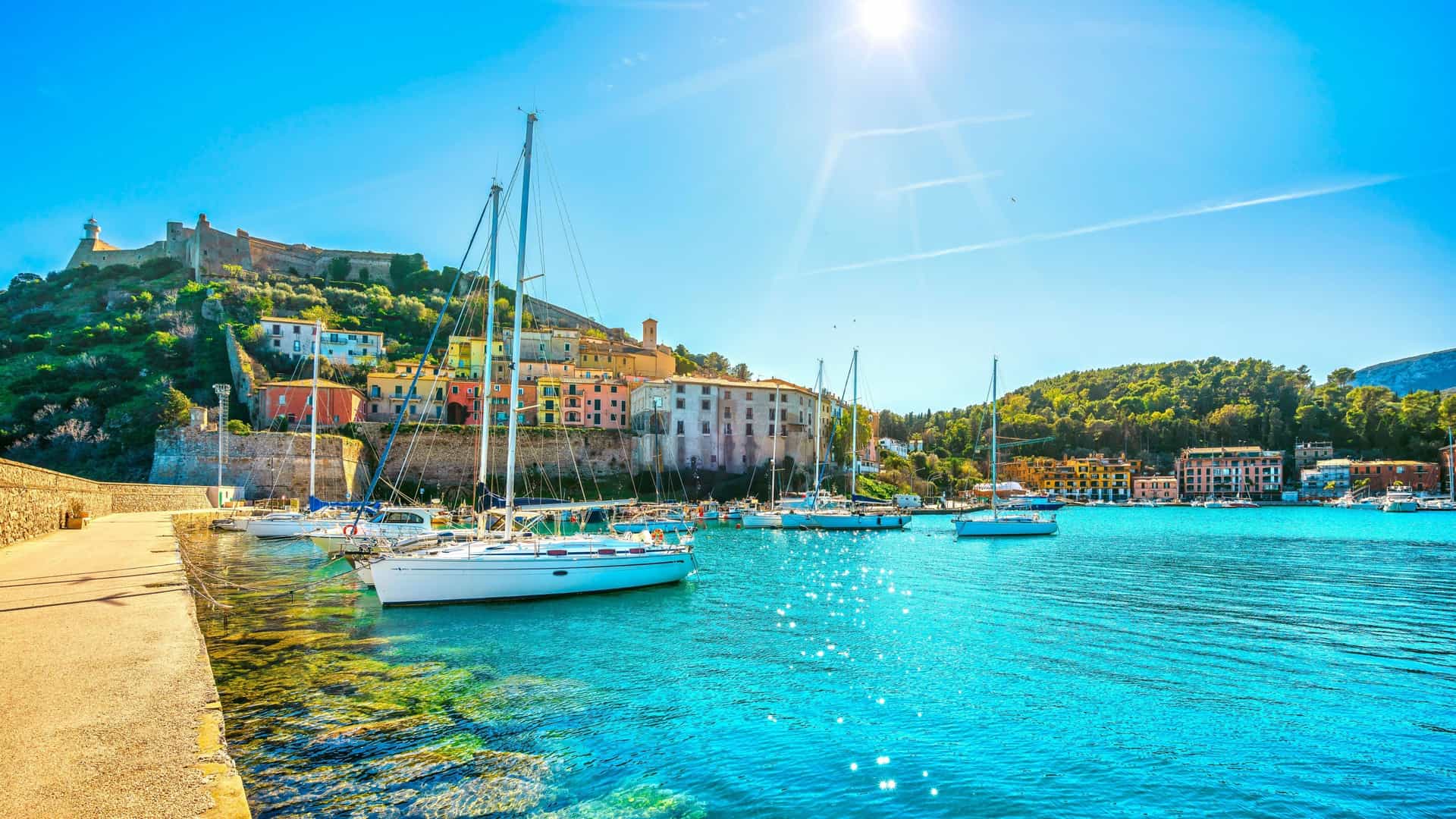 A scenic view of Porto Ercole, Italy, showing the vibrant blue sea with moored sailboats, colorful houses built on the hillside, and a historic fortress in the background.