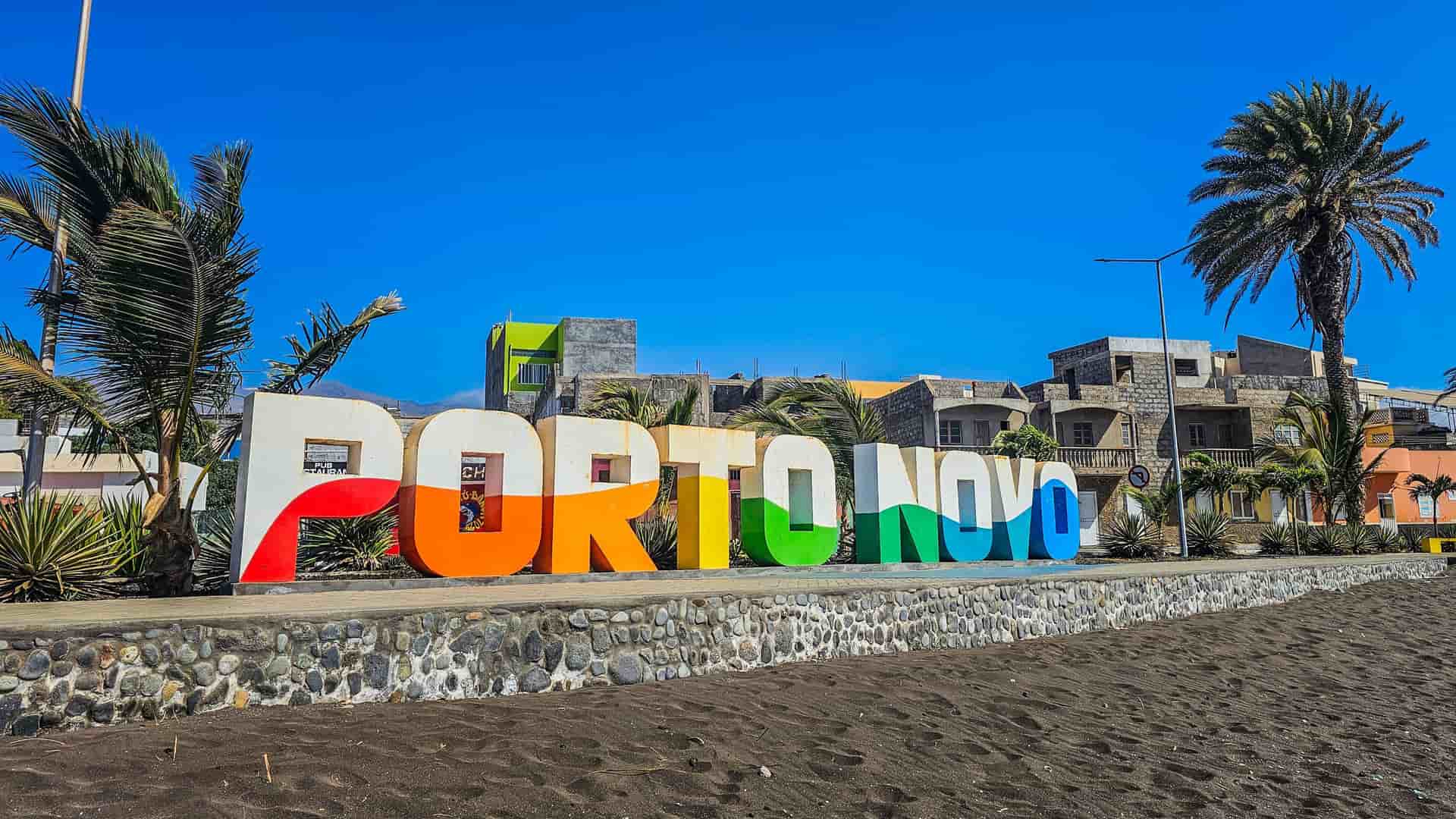 A large, colorful "PORTO NOVO" sign stands prominently on the beachfront, framed by palm trees and buildings, welcoming visitors to the city in Cape Verde.