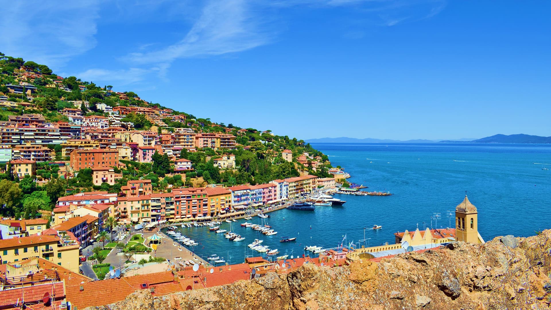 A panoramic view of Porto Santo Stefano, Italy, with its colorful houses built on a steep hillside overlooking the marina and the sparkling Tyrrhenian Sea.