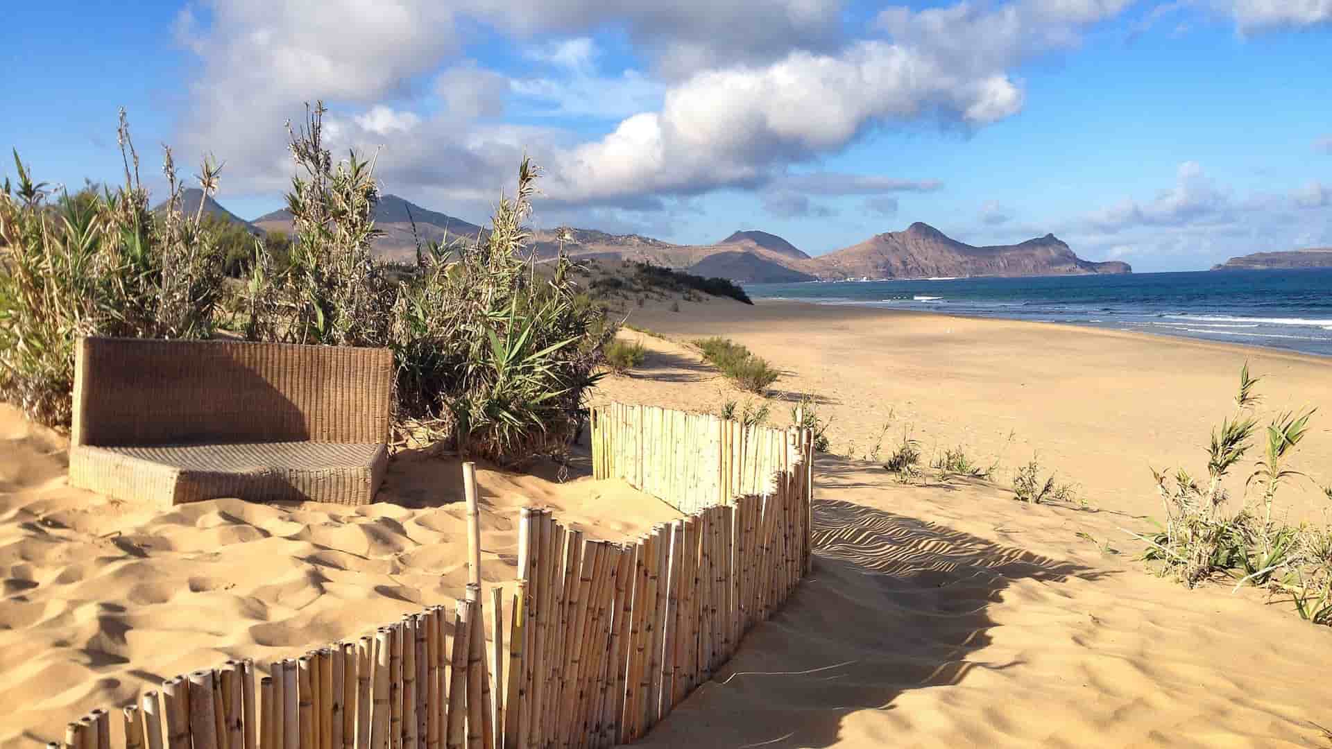 A beautiful Porto Santo beach landscape with a long stretch of golden sand, a wicker bench, a wooden fence, and the distinctive mountains of Madeira in the background.