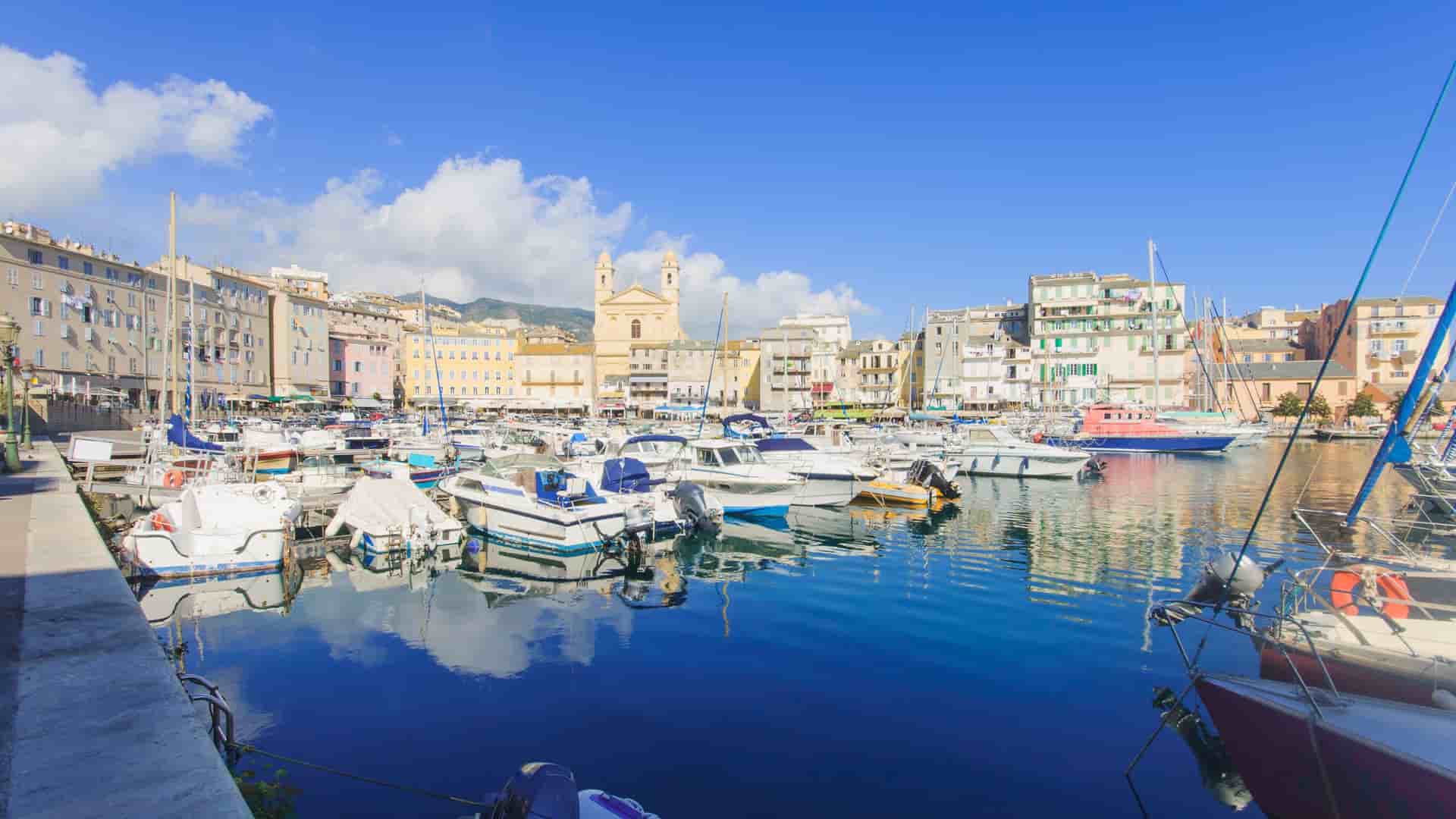 A scenic view of the charming Porto-Vecchio marina in Corsica, filled with boats and yachts and surrounded by a row of historic, multi-colored buildings under a clear blue sky.