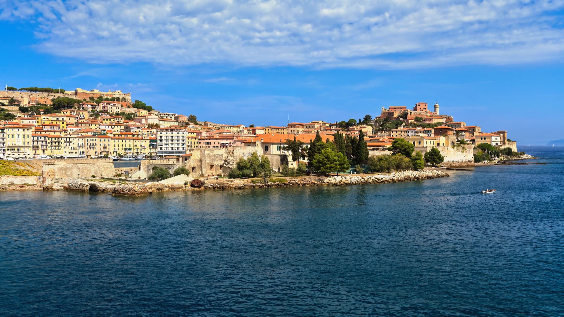 A beautiful wide shot of the Port of Portoferraio on Elba Island, Italy, showcasing the historic town's terracotta-roofed buildings and fortifications sprawling up the hillside from the blue sea.