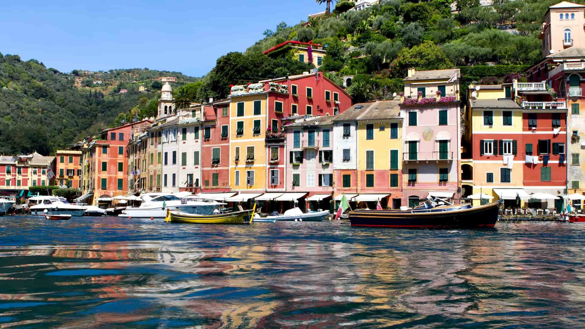 A classic view of the picturesque harbor of Portofino, Italy, with its iconic, brightly painted buildings reflected on the shimmering blue water and lush green hills in the background.