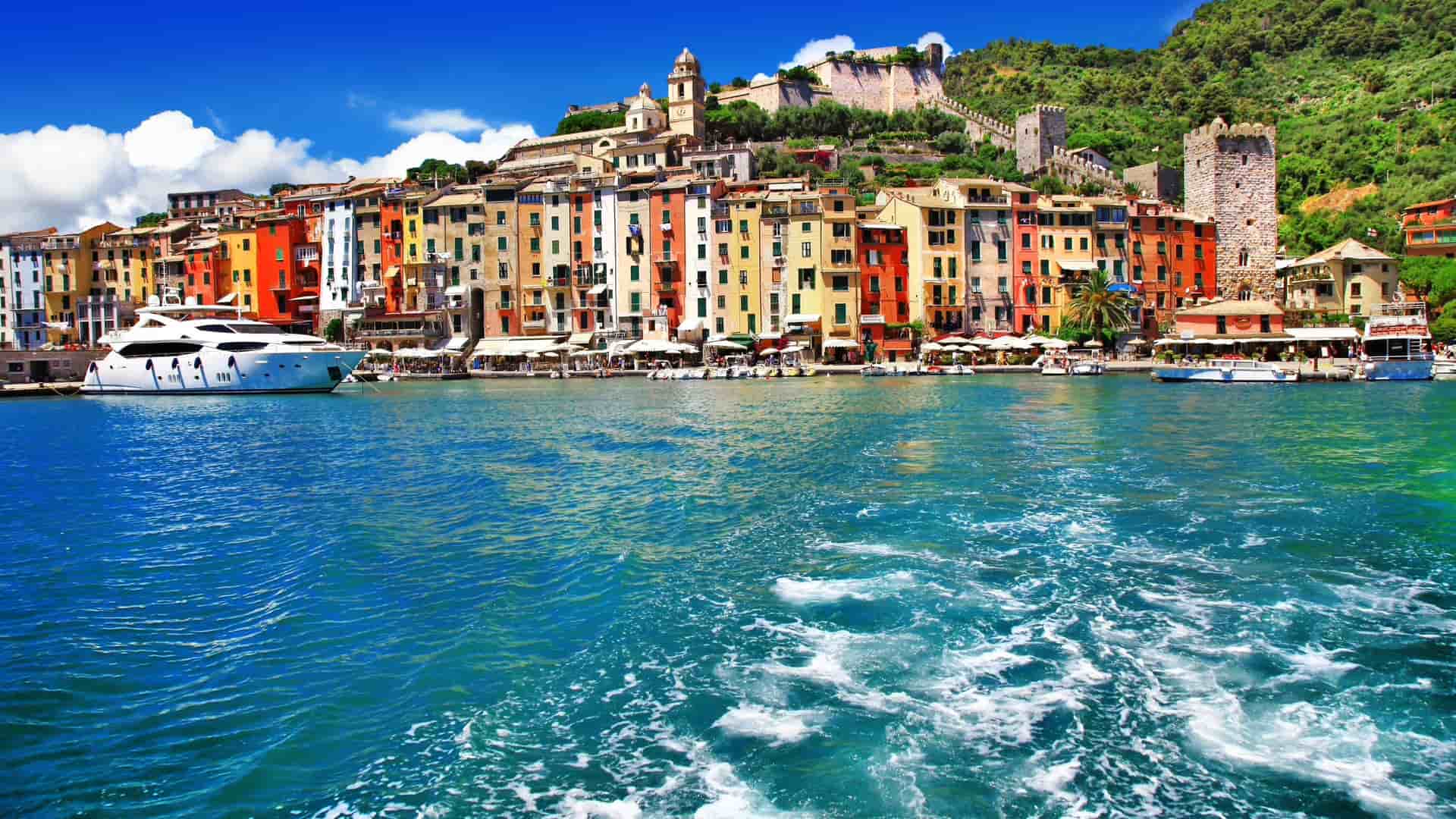 A scenic view of Portovenere, Italy, showcasing its charming harbor with colorful waterfront houses and a medieval castle on the hill under a bright blue sky.