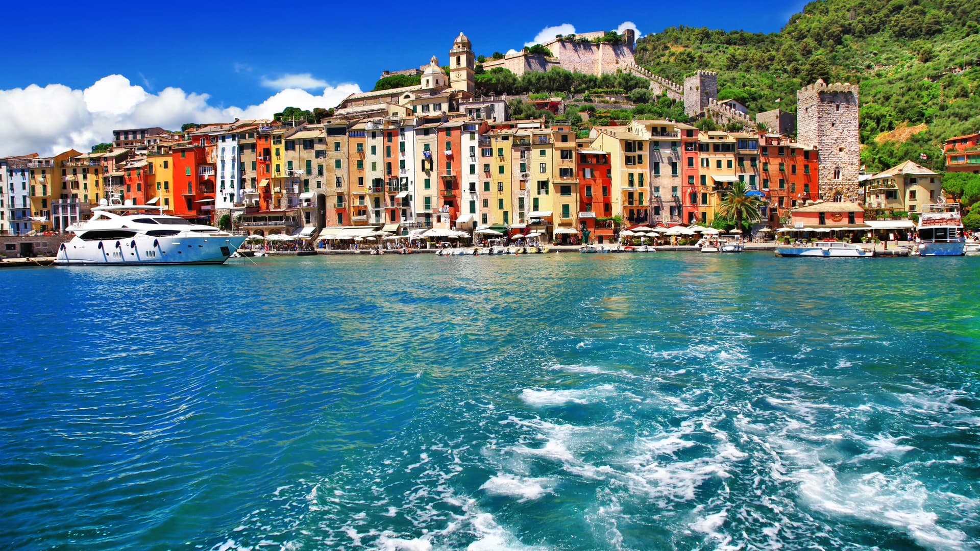 A scenic view of Portovenere, Italy, showcasing its charming harbor with colorful waterfront houses and a medieval castle on the hill under a bright blue sky.