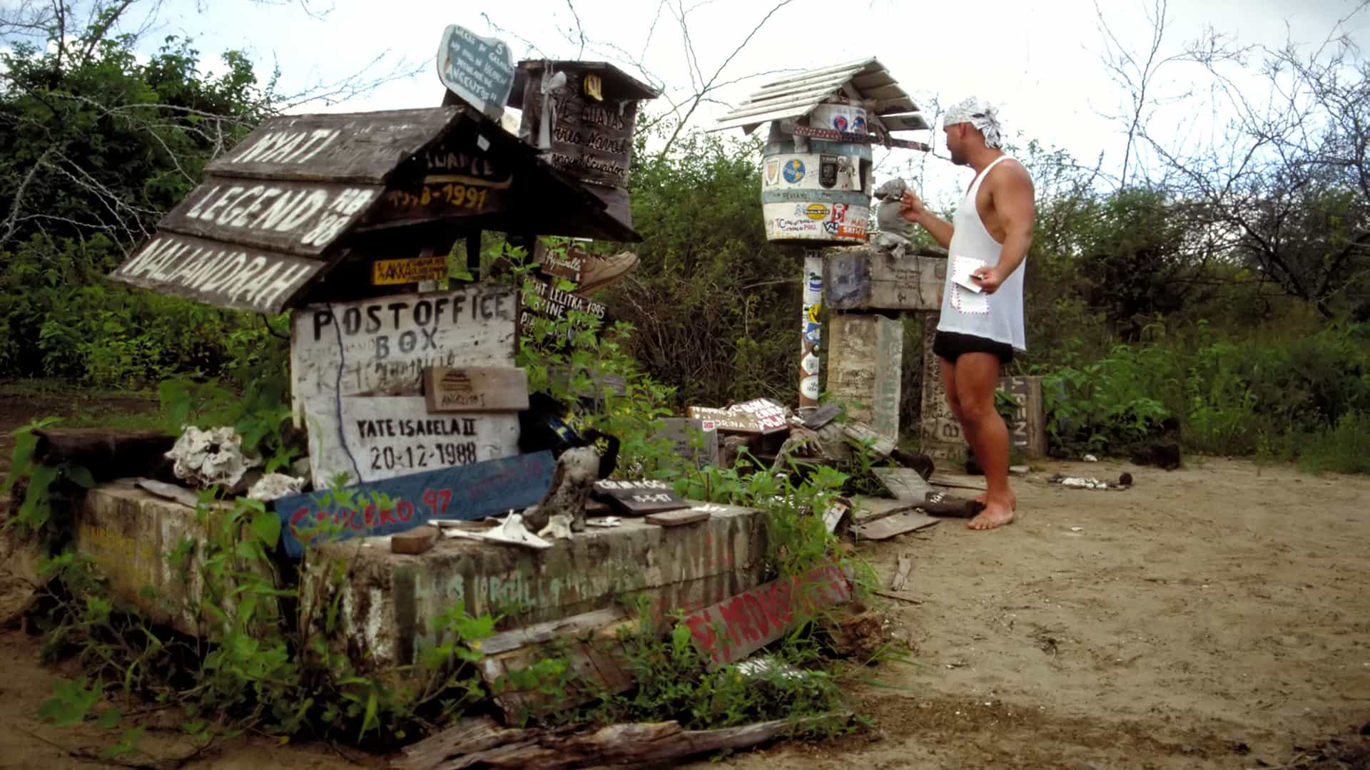 A barefoot man in a white tank top looks inside the historic Post Office Box on Floreana Island in the Galapagos, a rustic wooden landmark surrounded by weathered signs.