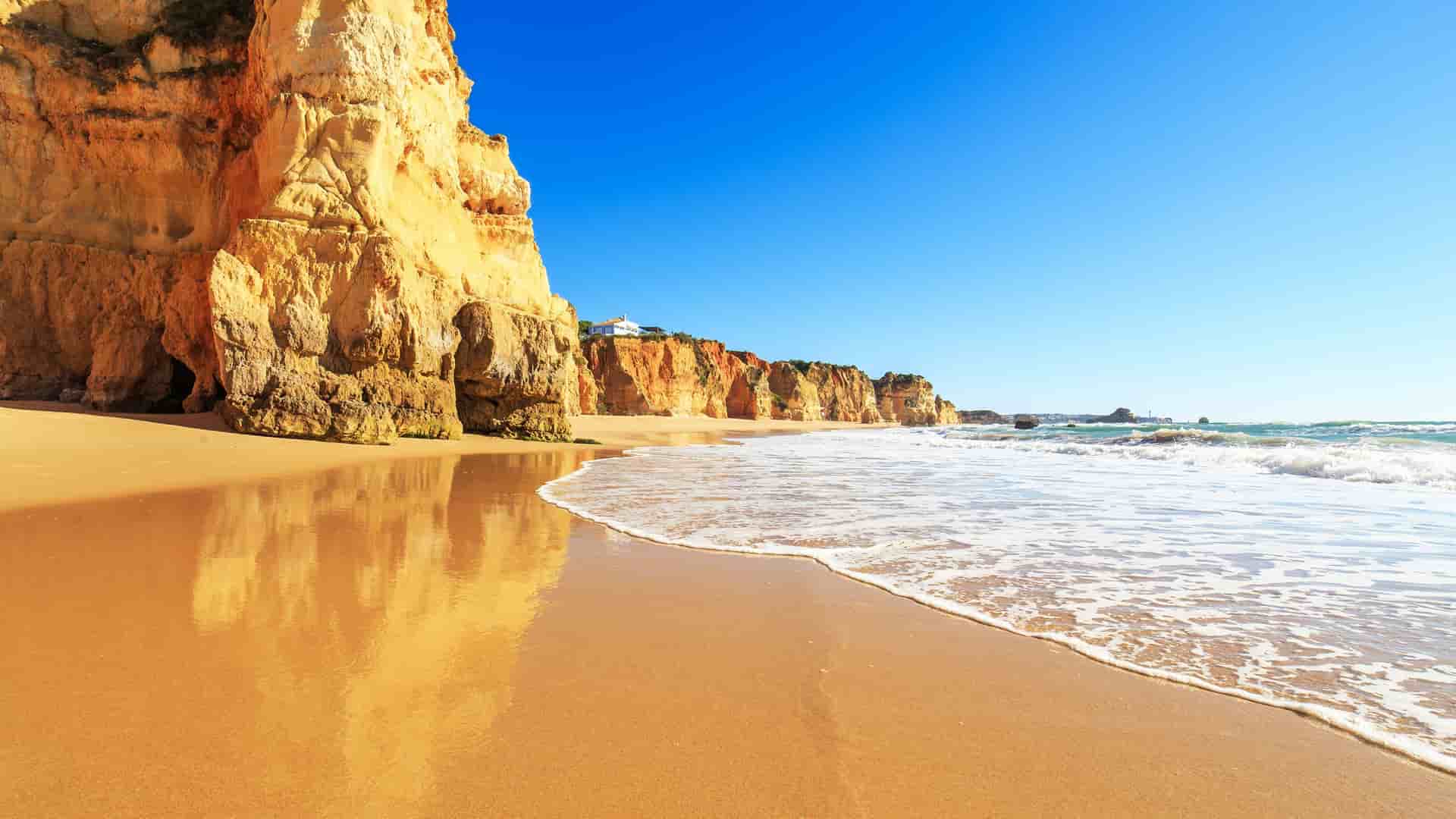 A sunny beach scene with golden sand and foamy waves, featuring large cliffs and rock formations along the coastline of Praia da Rocha in the Azores.