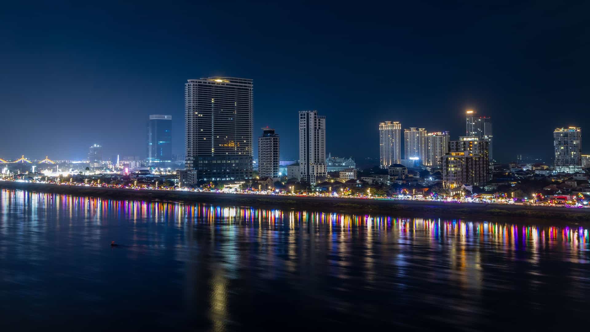 A wide-angle, long-exposure night shot of the illuminated city skyline of Prek Kdam, Cambodia, with vibrant light reflections shimmering on the water.