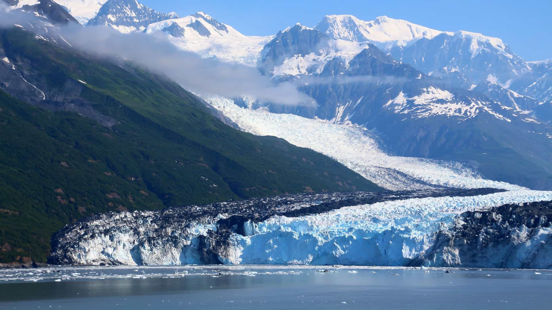 A wide-angle landscape of the beautiful Harvard Glacier in Prince William Sound, Alaska, with its stunning blue ice front and massive snow-covered mountains in the background.