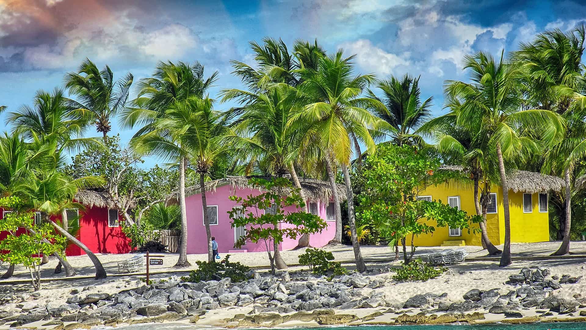 A scenic view of Princess Cays, Bahamas, featuring a row of colorful cabanas with thatched roofs, surrounded by swaying palm trees on a white-sand beach.