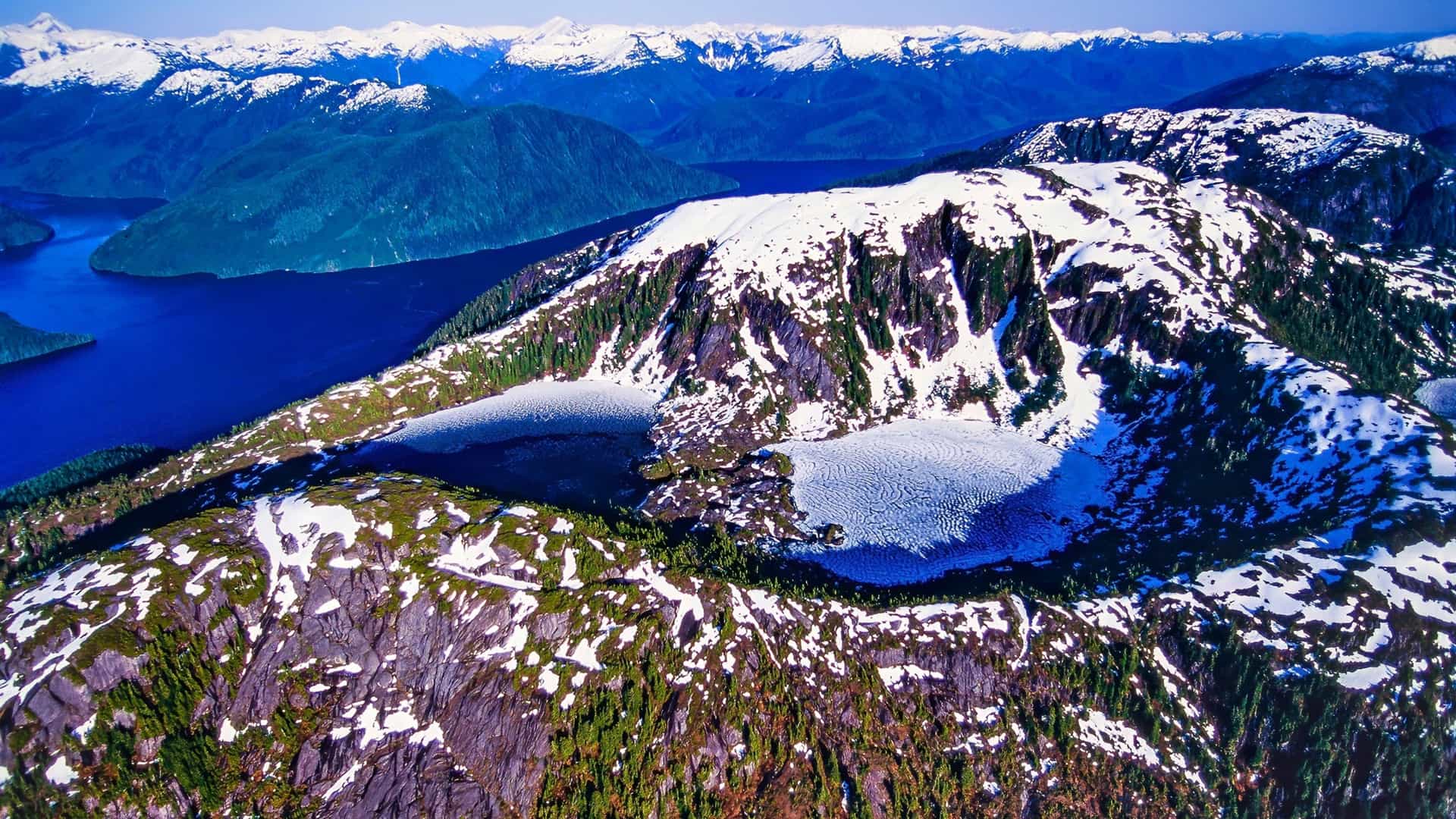 An aerial view of the snow-covered mountains of Princess Royal Island, Canada, with a partially frozen lake in the foreground and the beautiful blue ocean in the distance.