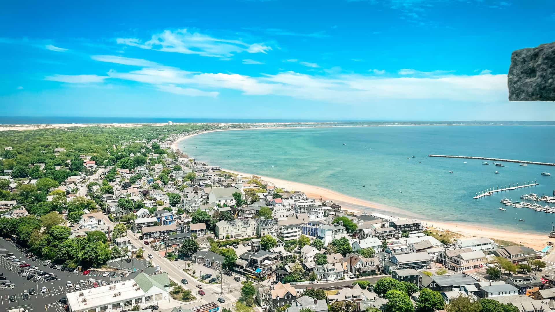 An aerial view of Provincetown, Massachusetts, showing the town's dense collection of houses and buildings curving along a long, sandy beach. The tranquil, turquoise water of the bay is filled with many boats, and a long sand spit extends into the distance under a bright, clear blue sky.