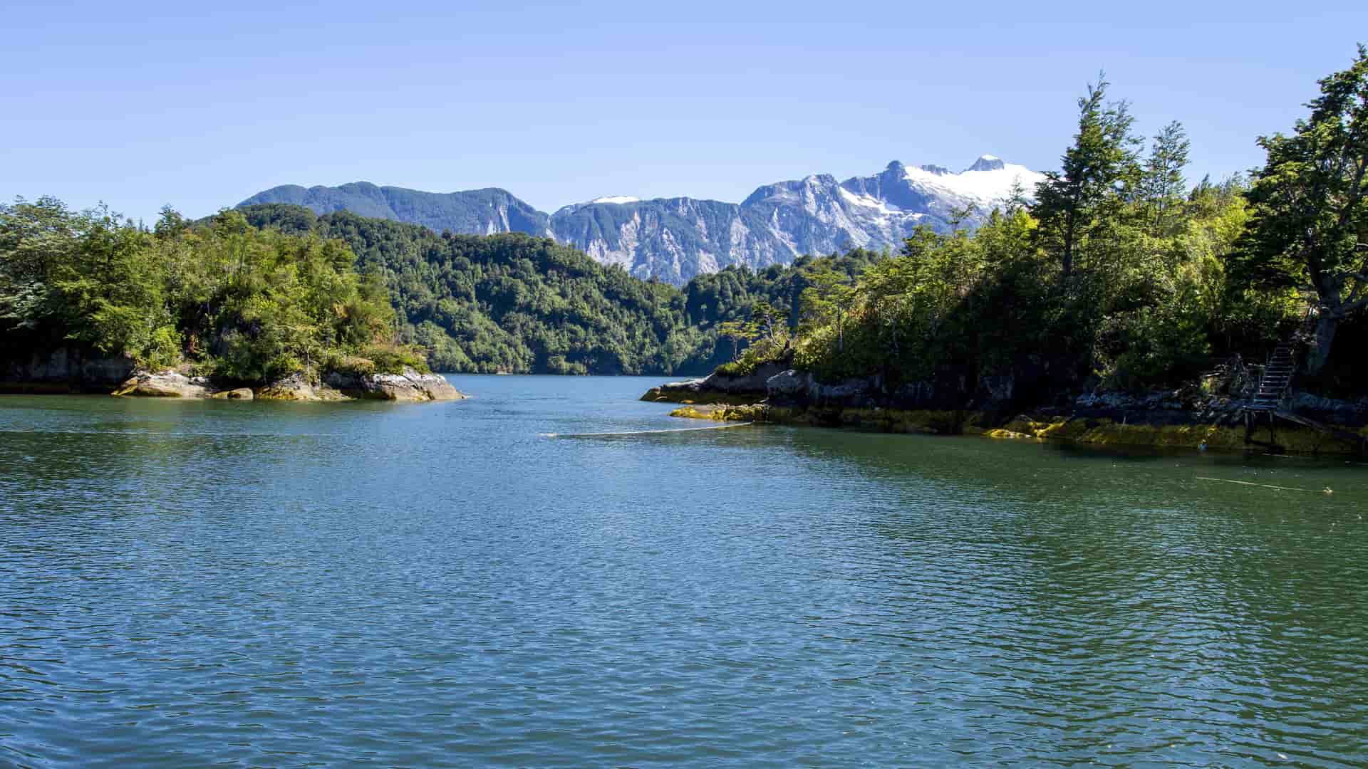 A stunning view of the Patagonian fjords near Puerto Chacabuco, Chile, featuring calm blue water, lush green forested hills, and snow-capped mountains in the distance.