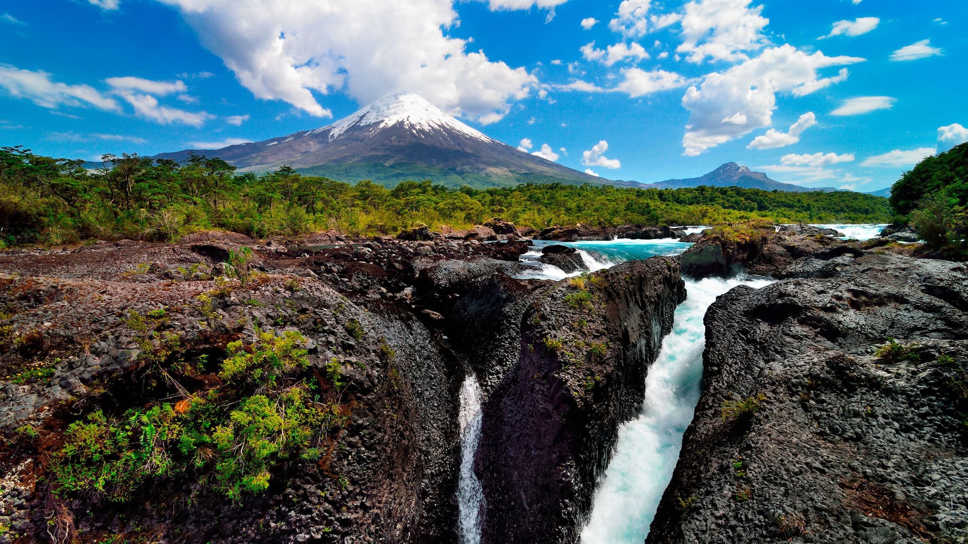 A wide shot of the spectacular Petrohué Falls near Puerto Montt, Chile, with turquoise water cascading over volcanic rock, set against the backdrop of the snow-capped Osorno Volcano.