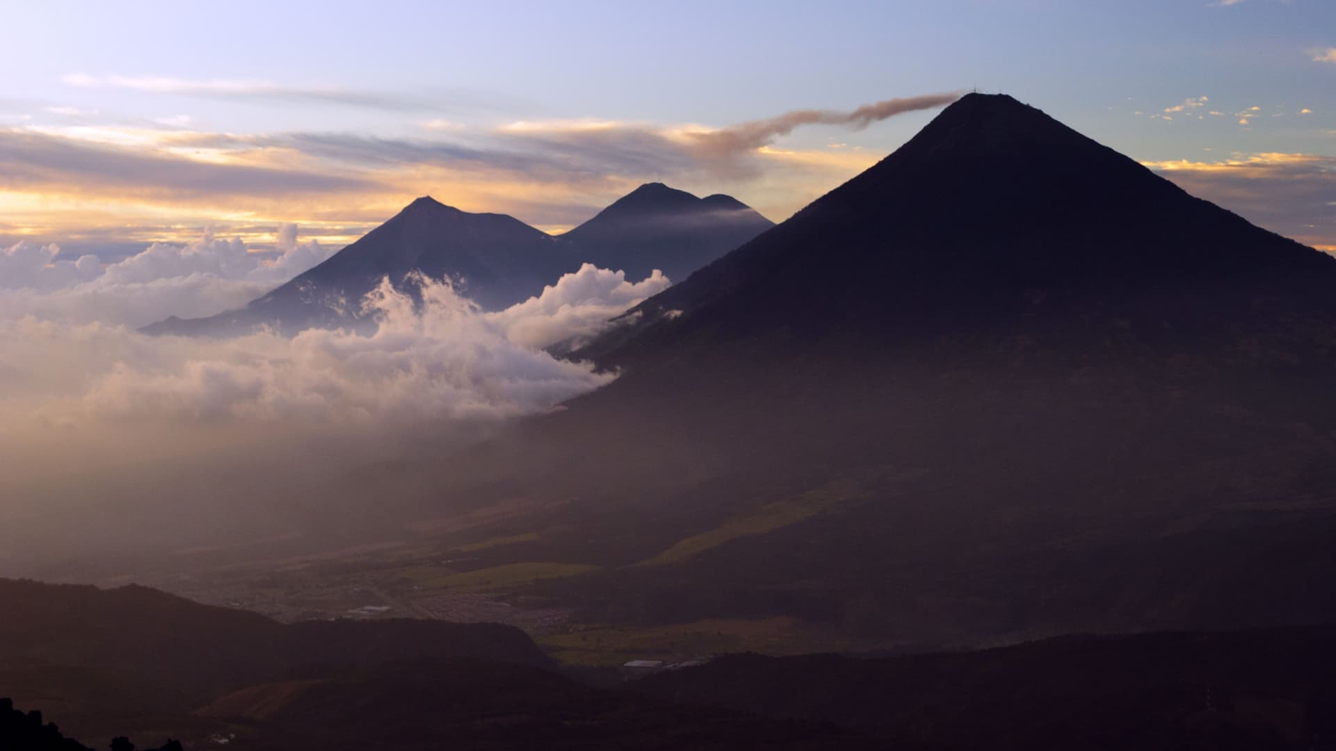 A wide-angle landscape shot of the three iconic volcanoes, Agua, Fuego, and Acatenango, in Guatemala, rising above the clouds during a vibrant sunrise, a scenic view near Puerto Quetzal.