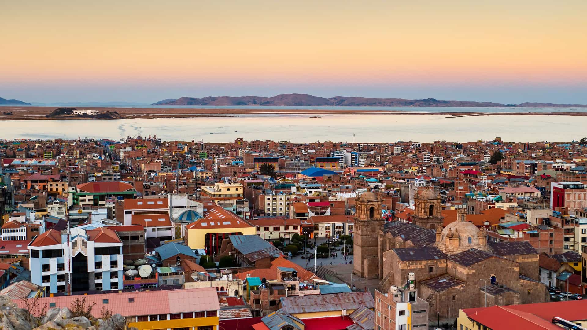 A panoramic, elevated view of the city of Puno, Peru, with its dense collection of buildings and a prominent, historic church with twin towers and a dome in the foreground. The city is situated on the shore of a vast, calm body of water, with distant hills visible on the far side and a colorful sunset sky above.