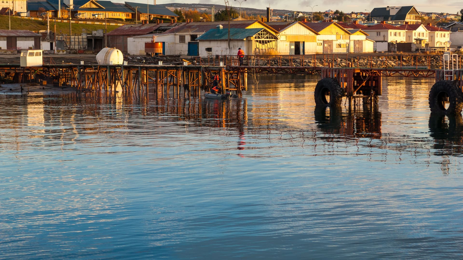 A serene waterside view of the historic wooden pier and colorful buildings lining the shore of Punta Arenas, Chile, during a calm and beautiful sunrise.