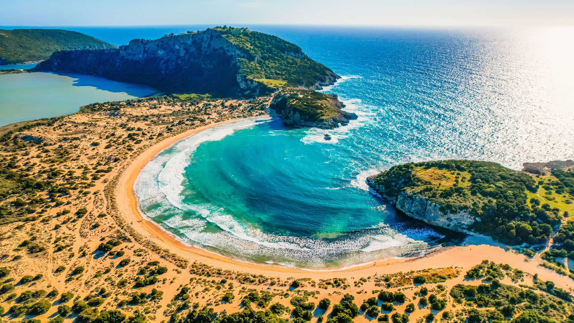 An aerial view of the famous omega-shaped Voidokilia Beach in Pylos, Greece, with its golden crescent of sand and a striking turquoise bay. A large, calm lagoon is visible on the left, separated from the open sea by a rocky peninsula, while the open ocean stretches to the right.