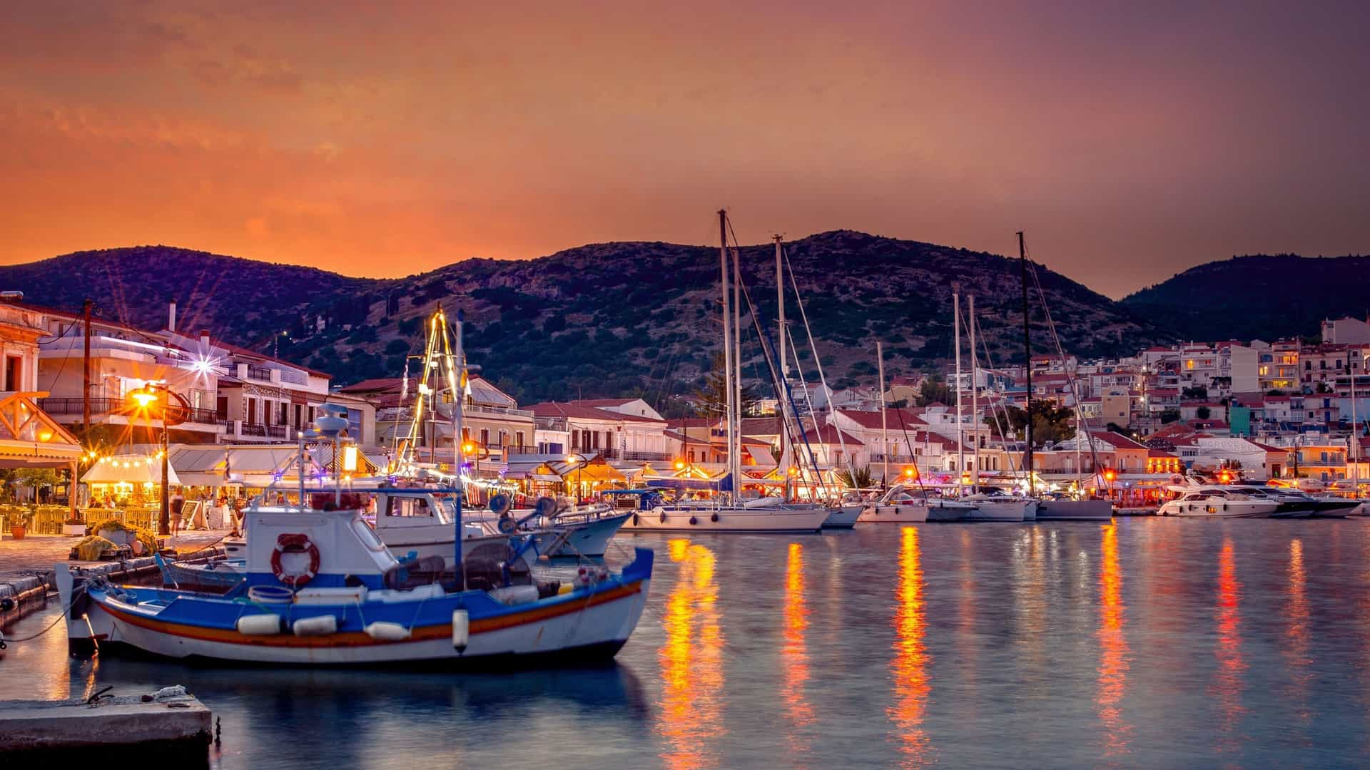A breathtaking sunset shot of the picturesque harbor in Pythagoreion, Samos, Greece, with fishing boats and sailboats docked in the calm water, and a backdrop of traditional buildings and mountains.