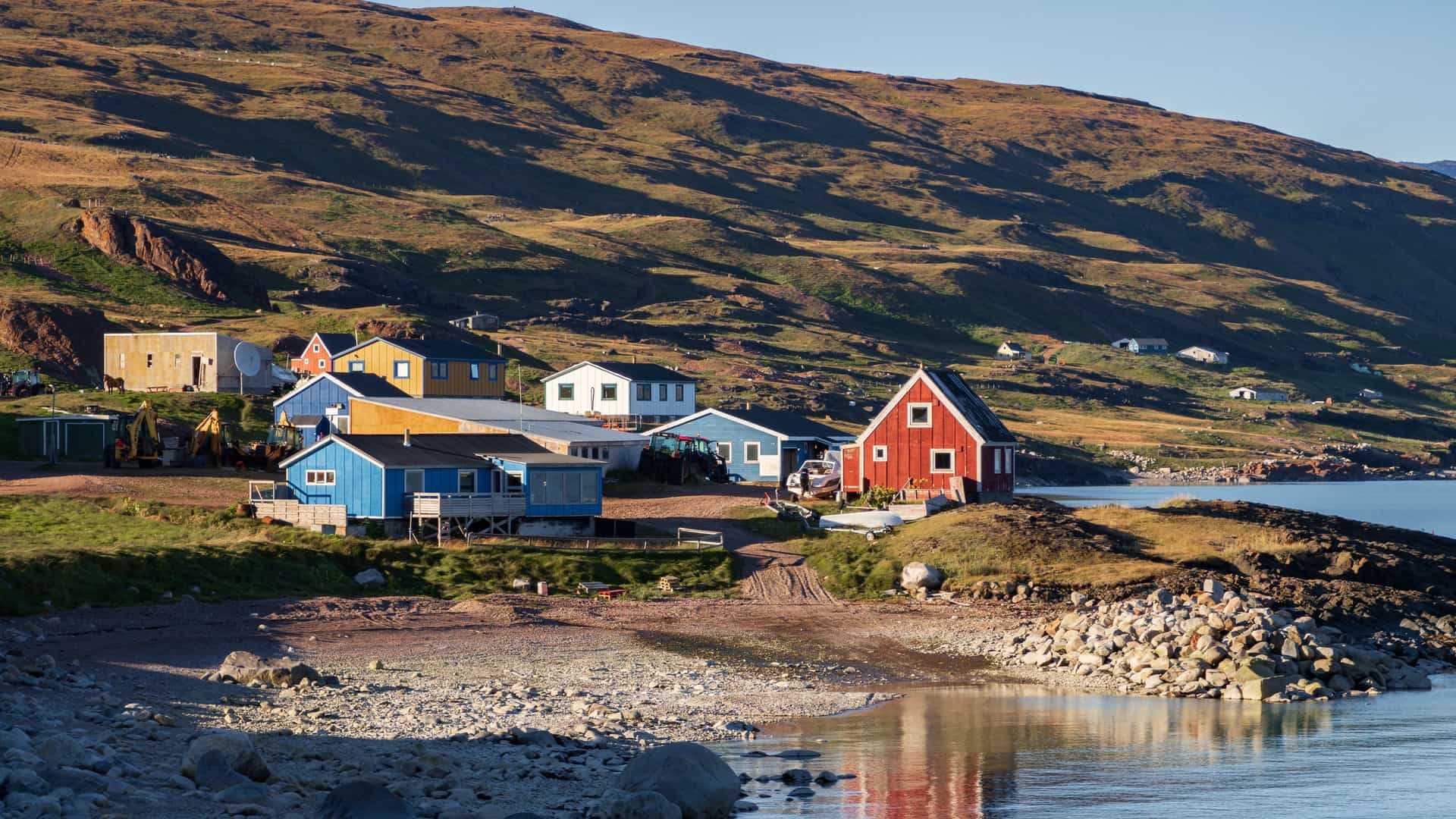 A scenic view of the village of Qassiarsuk in Greenland, with a cluster of colorful houses—including a prominent red one—perched on a rocky shoreline. The settlement is set against the backdrop of a vast, textured, brown and green grassy hillside, with a calm body of water in the foreground.
