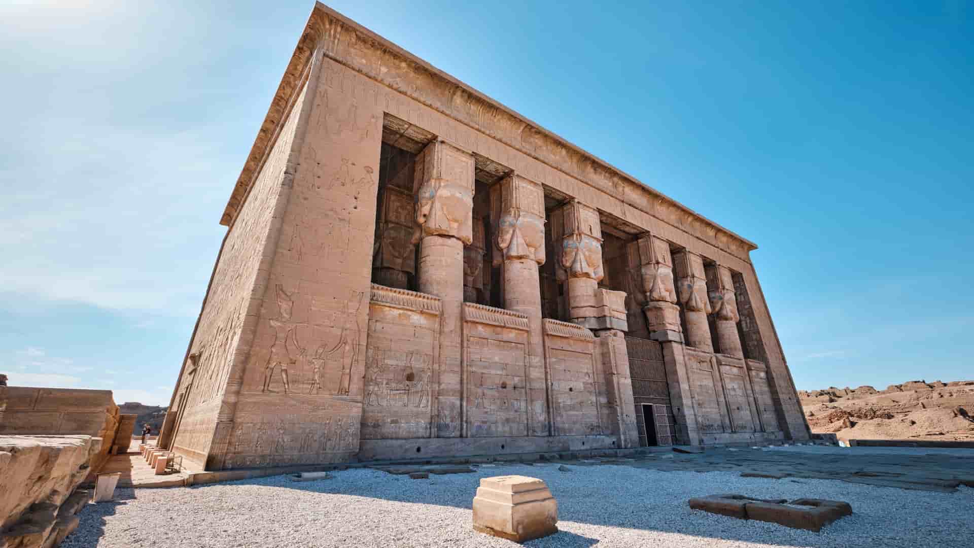 A low-angle shot of the monumental stone facade of the Temple of Hathor in Dendera, Egypt, near the city of Qena, with its intricately carved columns and hieroglyphics.