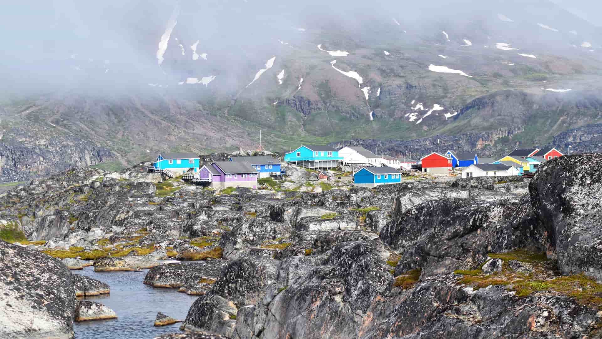 Brightly colored houses sit on a rugged, rocky black landscape with snow-dusted mountains in the misty town of Qeqertarsuaq, Greenland.