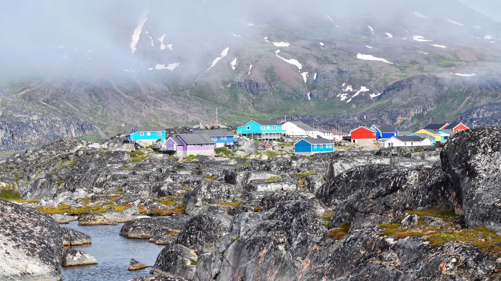 Brightly colored houses sit on a rugged, rocky black landscape with snow-dusted mountains in the misty town of Qeqertarsuaq, Greenland.