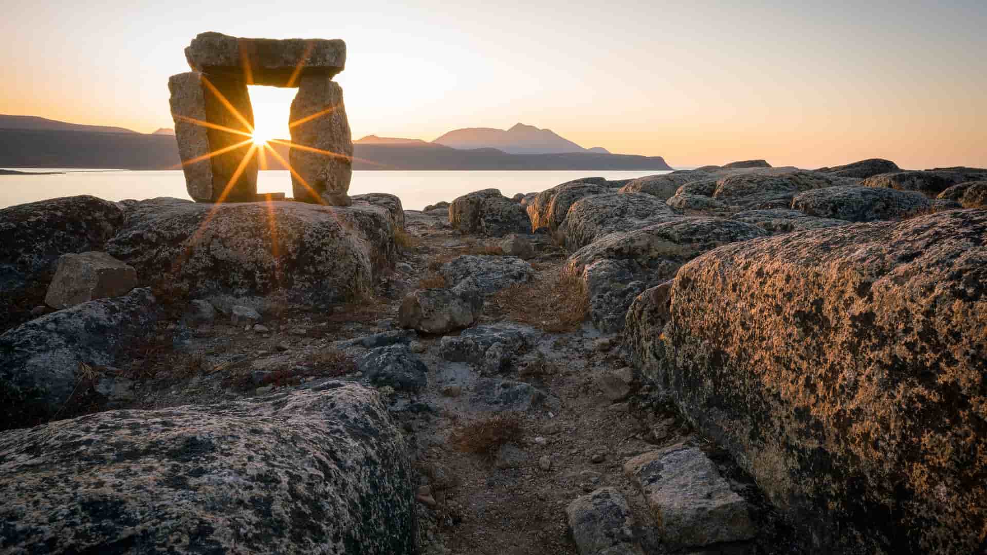 A scenic sunrise shot in the Arctic wilderness of Qikiqtarjuaq, Nunavut, with the sun's rays shining through a traditional Inuit inukshuk stone marker overlooking a tranquil ocean bay and mountains in the distance.