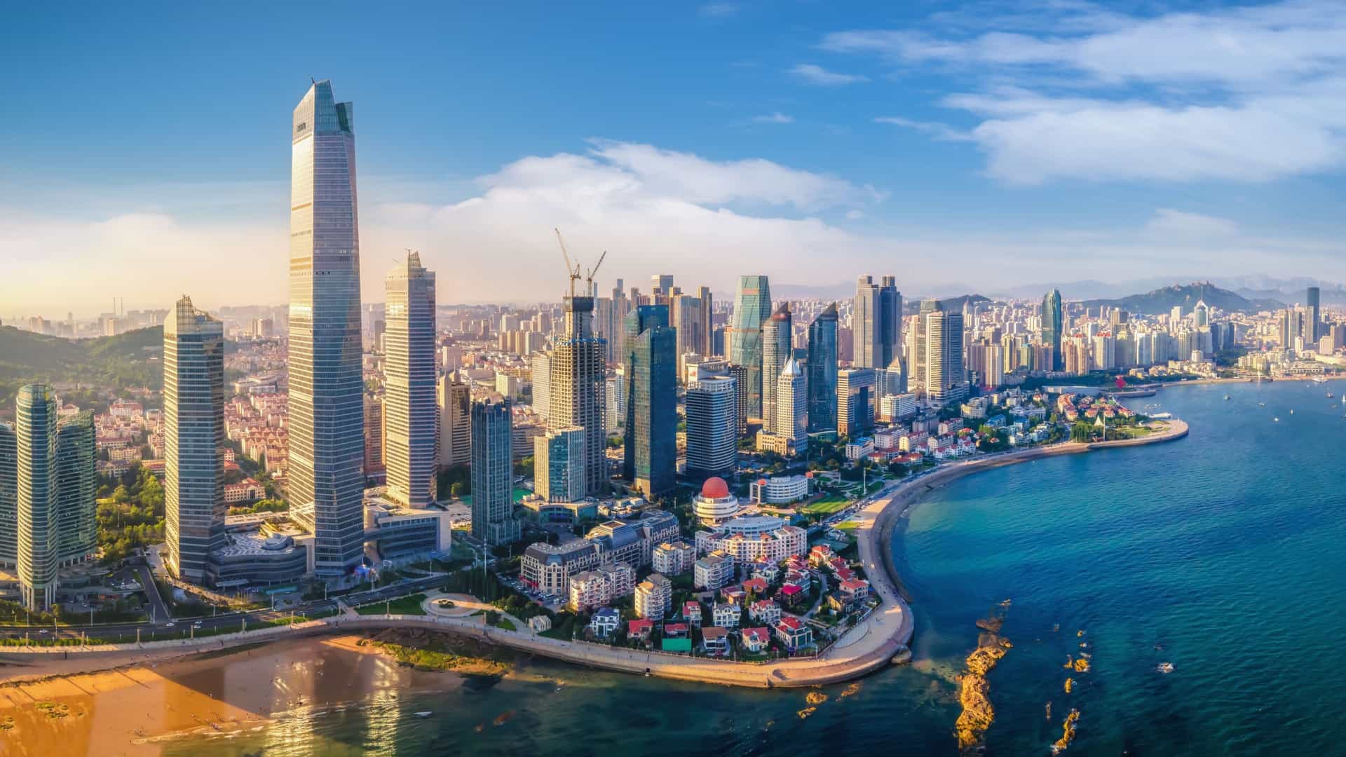 An aerial shot of the stunning modern skyline of Qingdao, China, with towering skyscrapers and buildings curving along the coast, highlighting the city's beautiful beachside location.